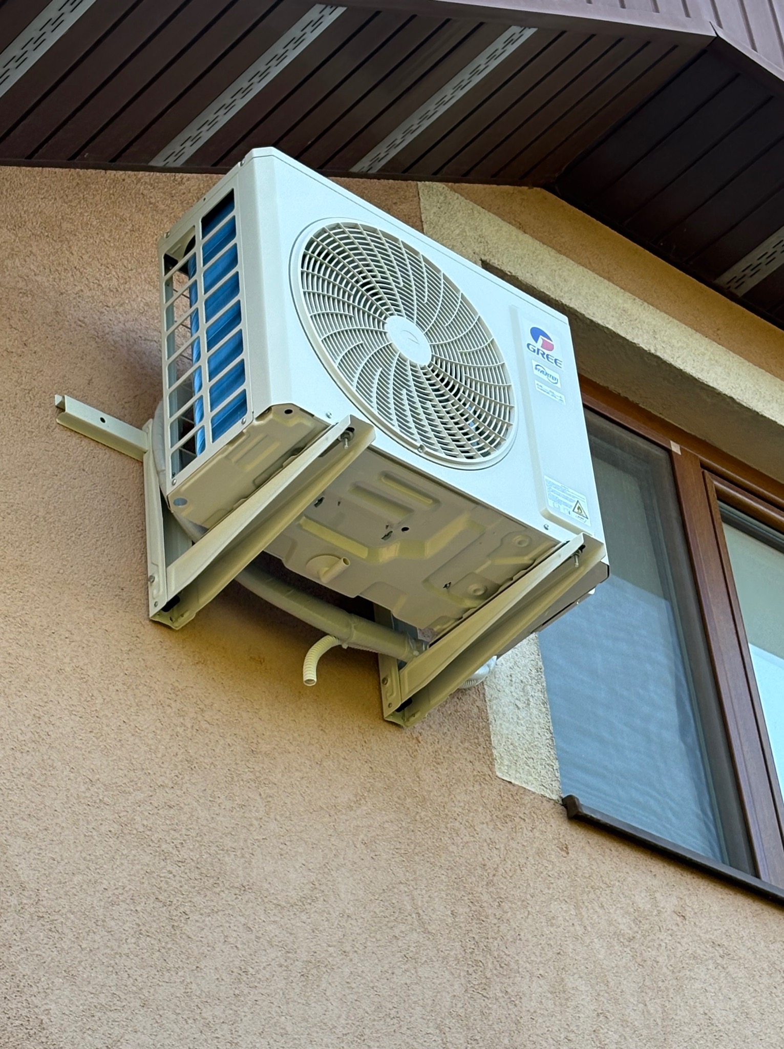 Biała klimatyzacja GREE zamontowana na elewacji budynku obok okna. Widoczne elementy montażowe i rurka odprowadzająca skropliny. Ujęcie z dołu.