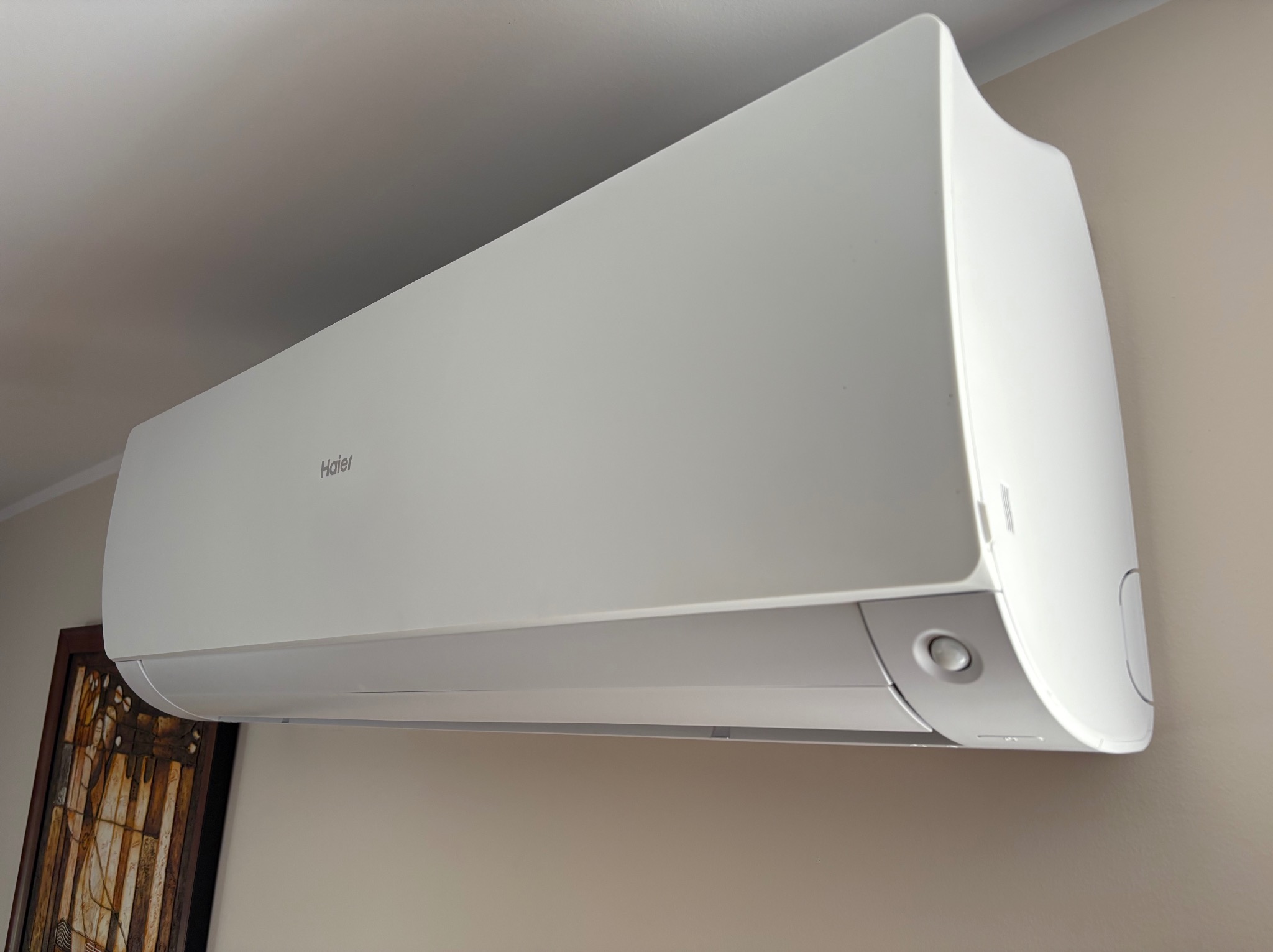 Biała, zamontowana klimatyzacja Haier na ścianie w pomieszczeniu, widoczny fragment obrazu w ramie, oświetlenie dzienne, ujęcie z dołu.