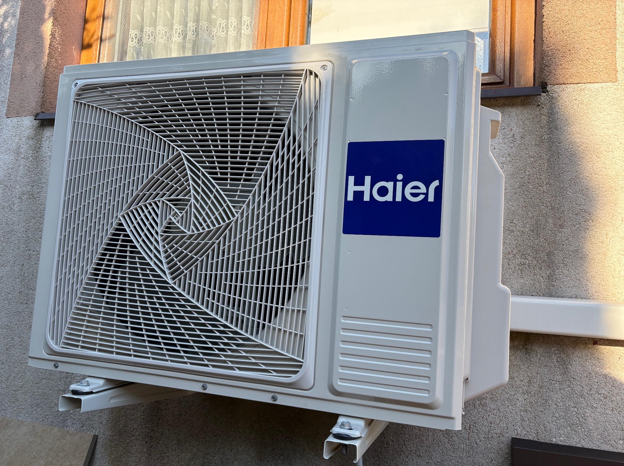 Biały klimatyzator Haier zamontowany na zewnątrz budynku, widoczne detale montażu i fragment okna z firanką w tle. Ujęcie z dołu, podkreślające solidność instalacji.