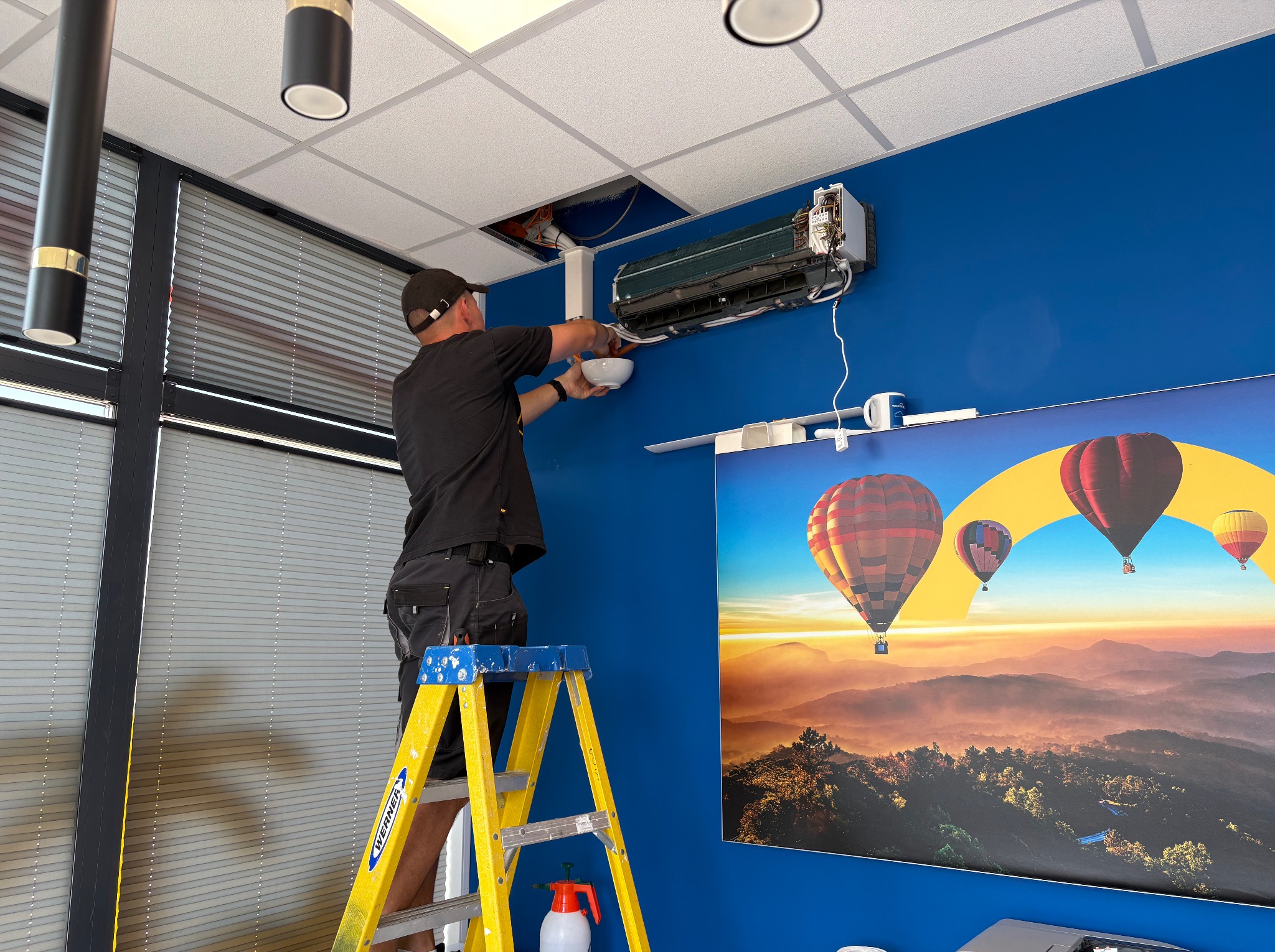 Serwisant na drabinie naprawia klimatyzator ścienny w biurze z niebieską ścianą i plakatem z balonami, widoczne narzędzia i otwarty sufit.