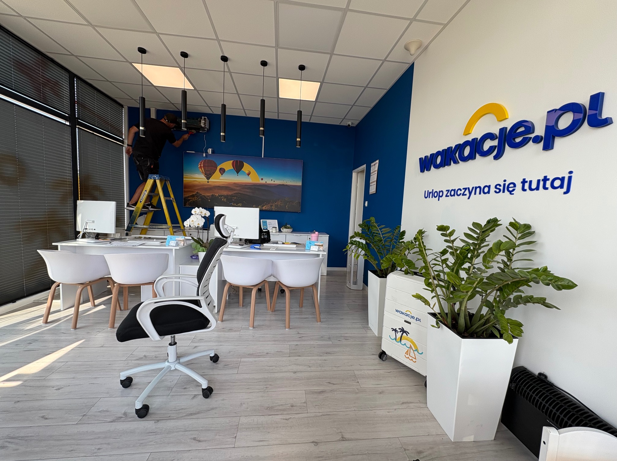 Montaż klimatyzacji w biurze podróży: mężczyzna na drabinie instaluje urządzenie na ścianie w nowoczesnym wnętrzu z logo firmy Wakacje.pl.