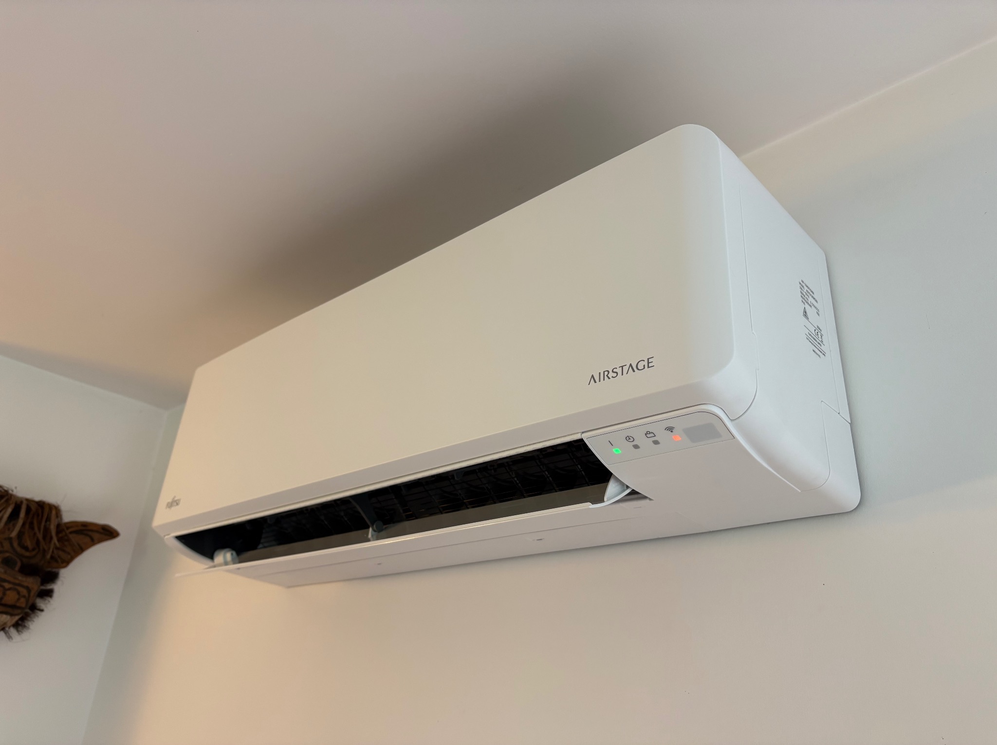 Biała klimatyzacja AIRSTAGE zamontowana na ścianie w Żukowie. Widoczne diody sygnalizacyjne i fragment dekoracji etnicznej po lewej stronie.