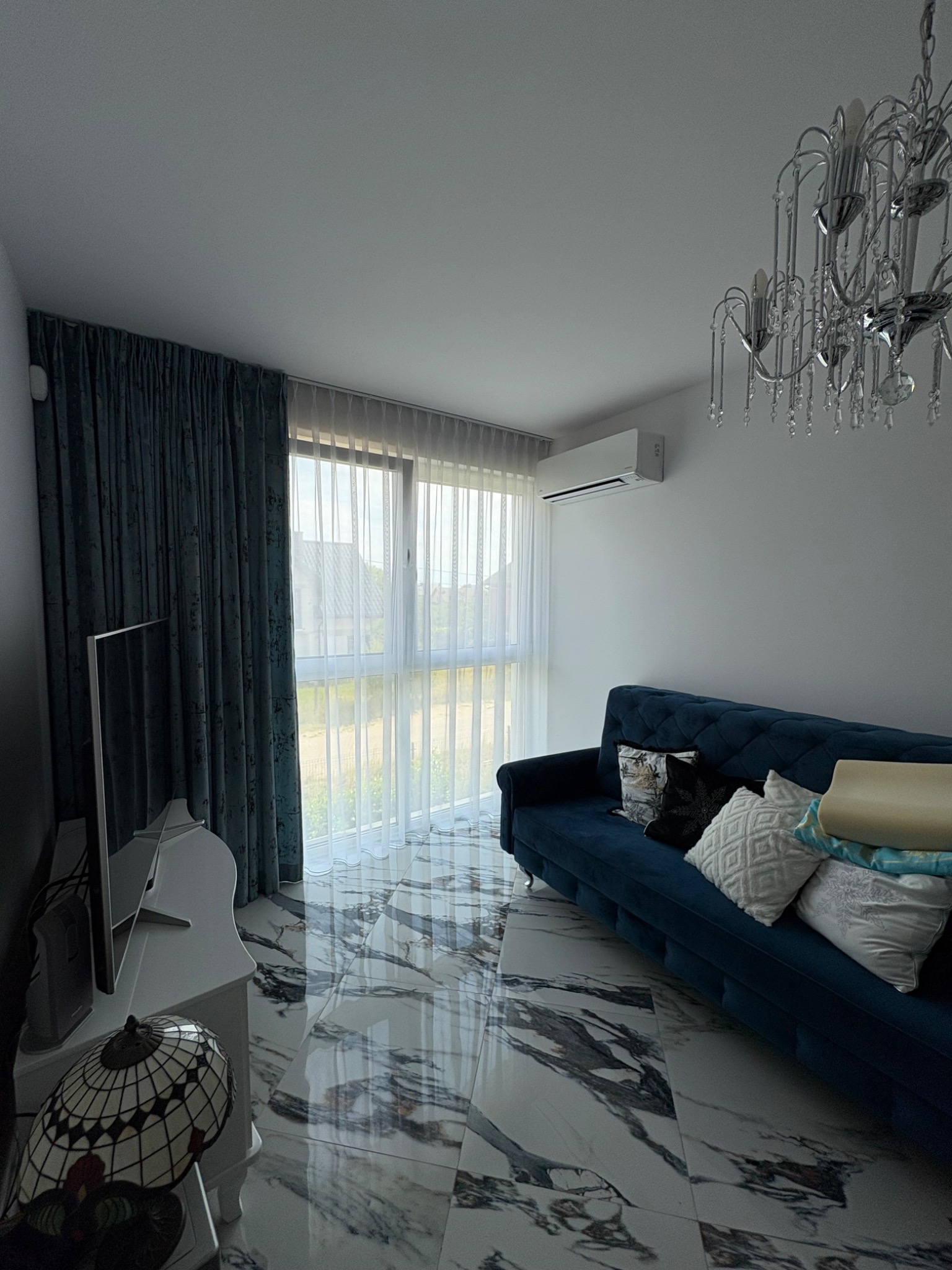 Elegancki salon z klimatyzacją ścienną, niebieską sofą i marmurową podłogą. Widoczne okna z firanami i zasłonami oraz ozdobny żyrandol.
