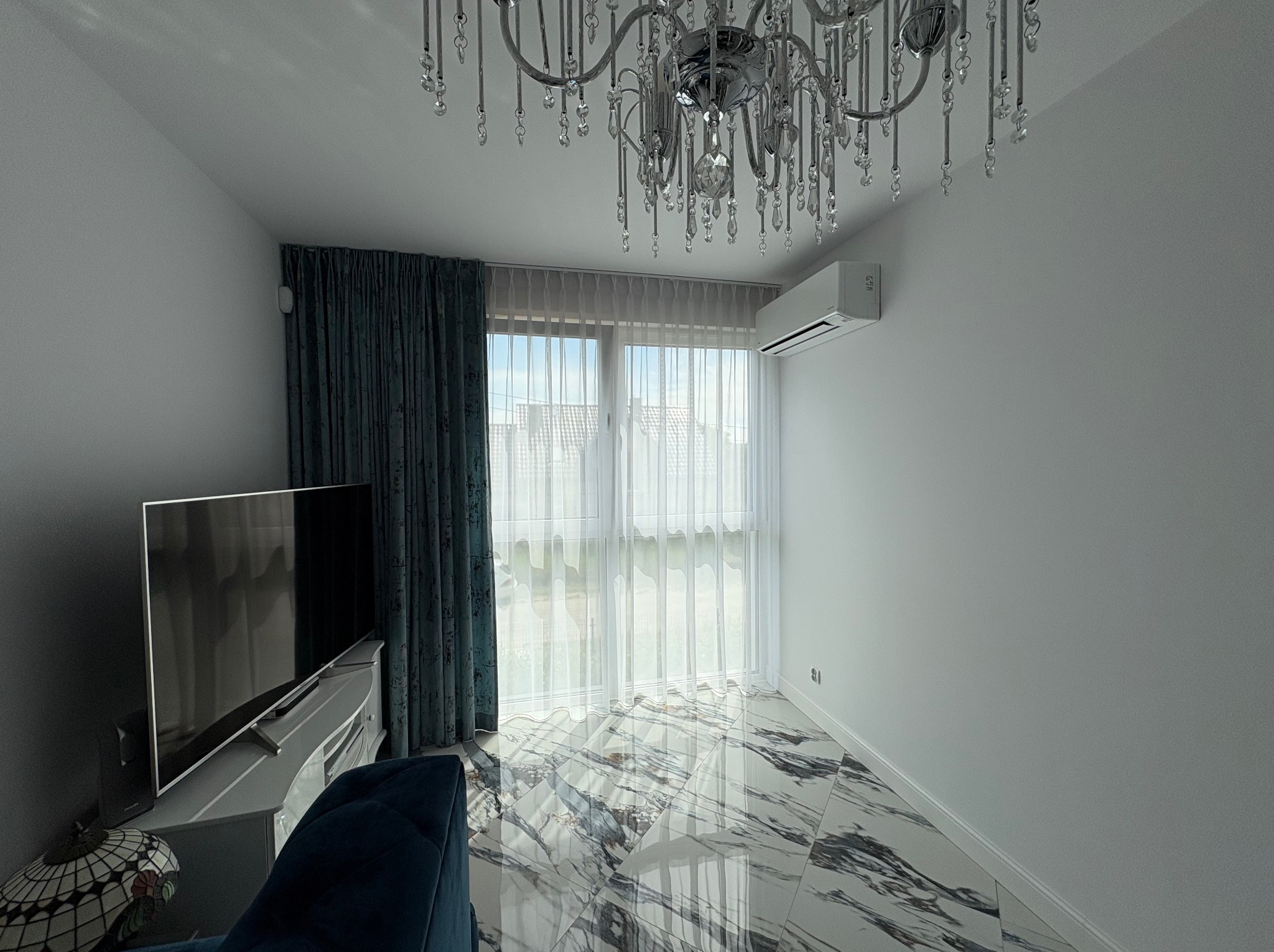 Nowoczesny salon z klimatyzatorem ściennym, kryształowym żyrandolem i marmurową podłogą. Widoczne zasłony i telewizor. Jasne, przestronne wnętrze.