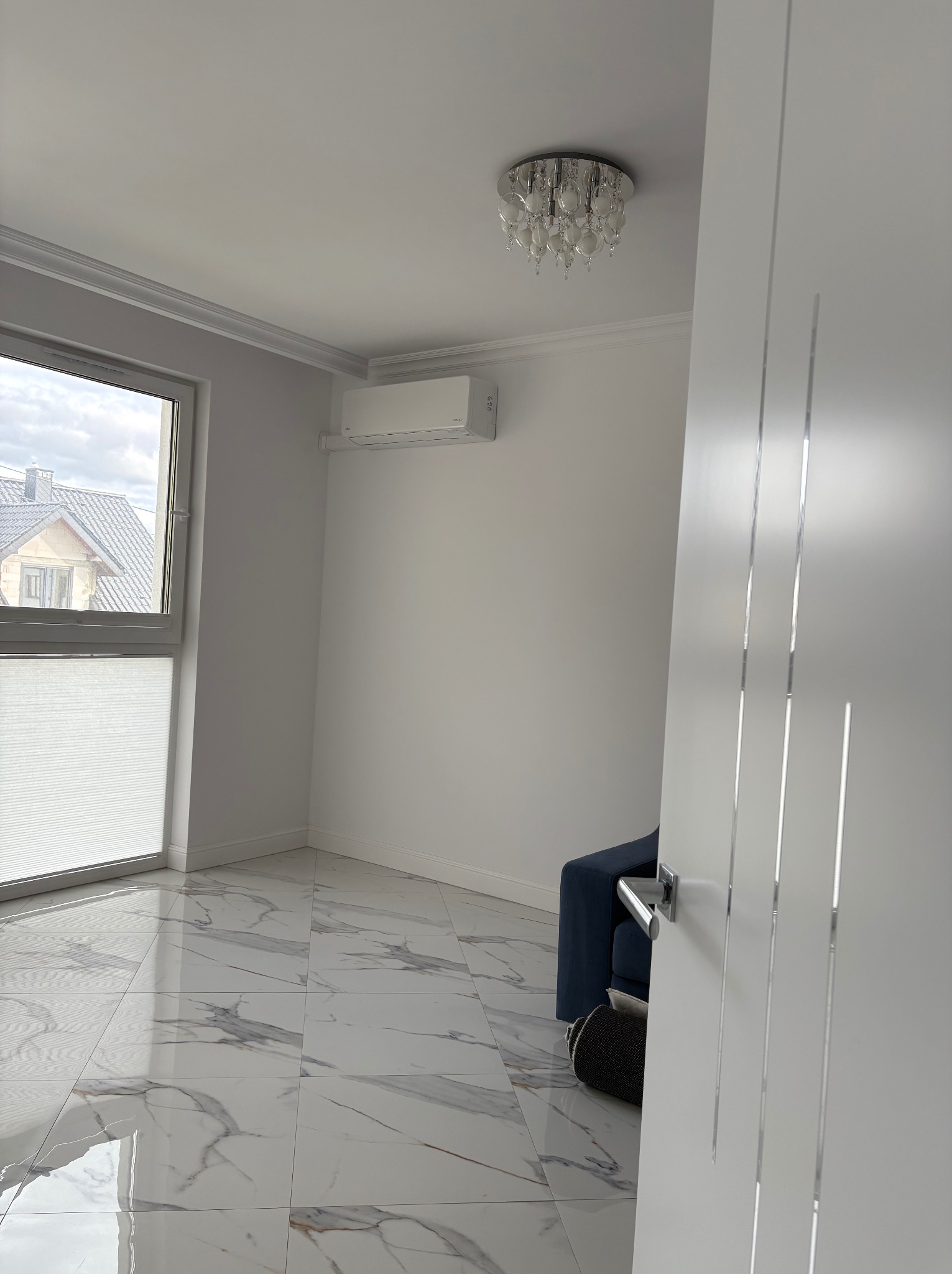 Nowoczesny pokój z klimatyzacją ścienną, marmurową podłogą i ozdobną lampą sufitową. Widok przez uchylone drzwi na minimalistyczne wnętrze.