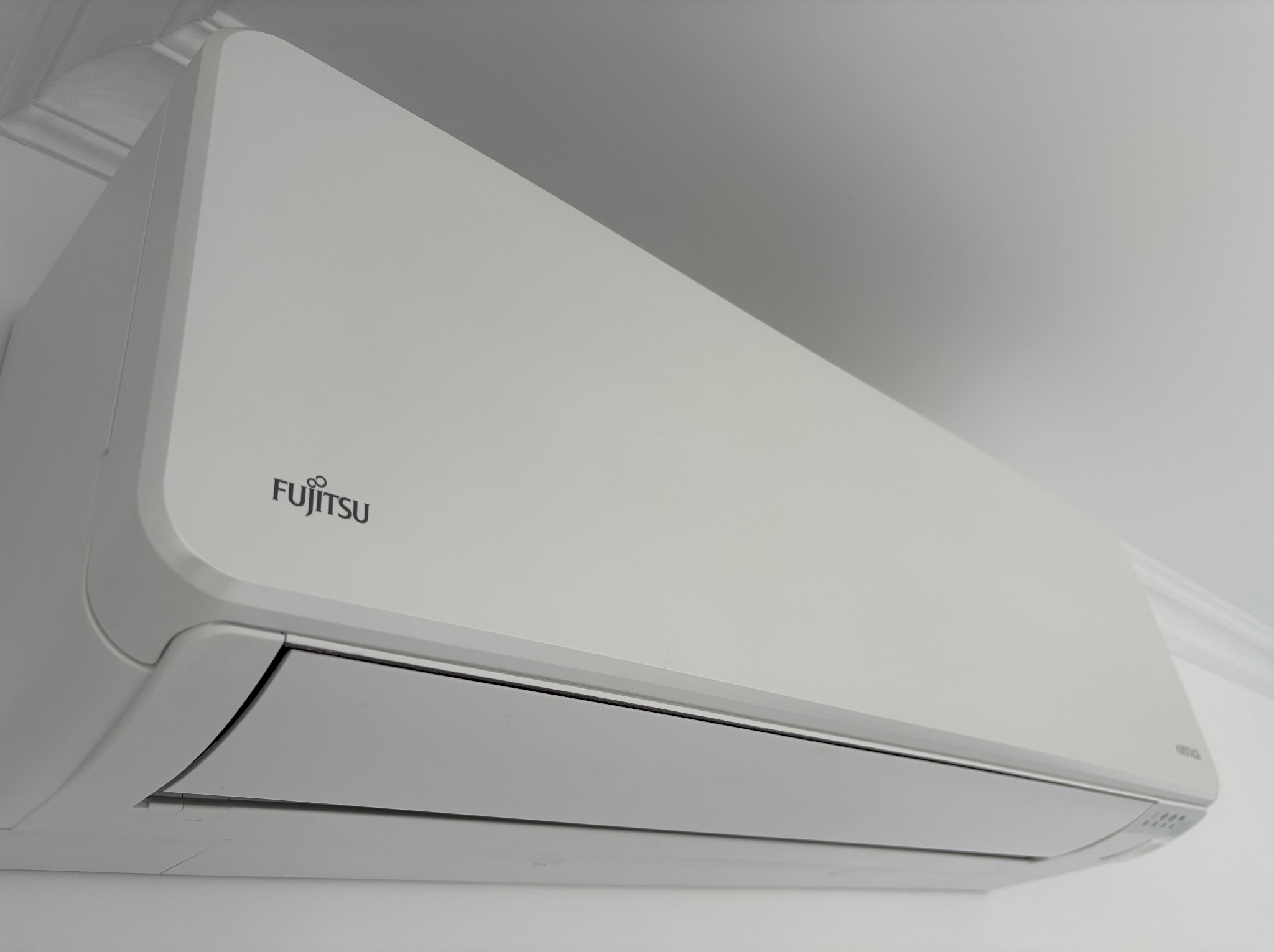 Biała jednostka wewnętrzna klimatyzacji Fujitsu zamontowana na ścianie, widoczna z perspektywy lekko od dołu, z detalami wykończenia sufitu w tle.
