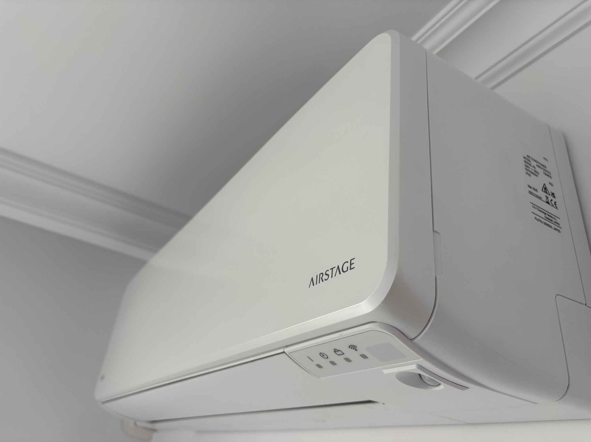 Biała klimatyzacja AIRSTAGE zamontowana na ścianie, widok z dołu pod kątem. Widoczne detale urządzenia i fragment sufitu z dekoracyjną listwą.