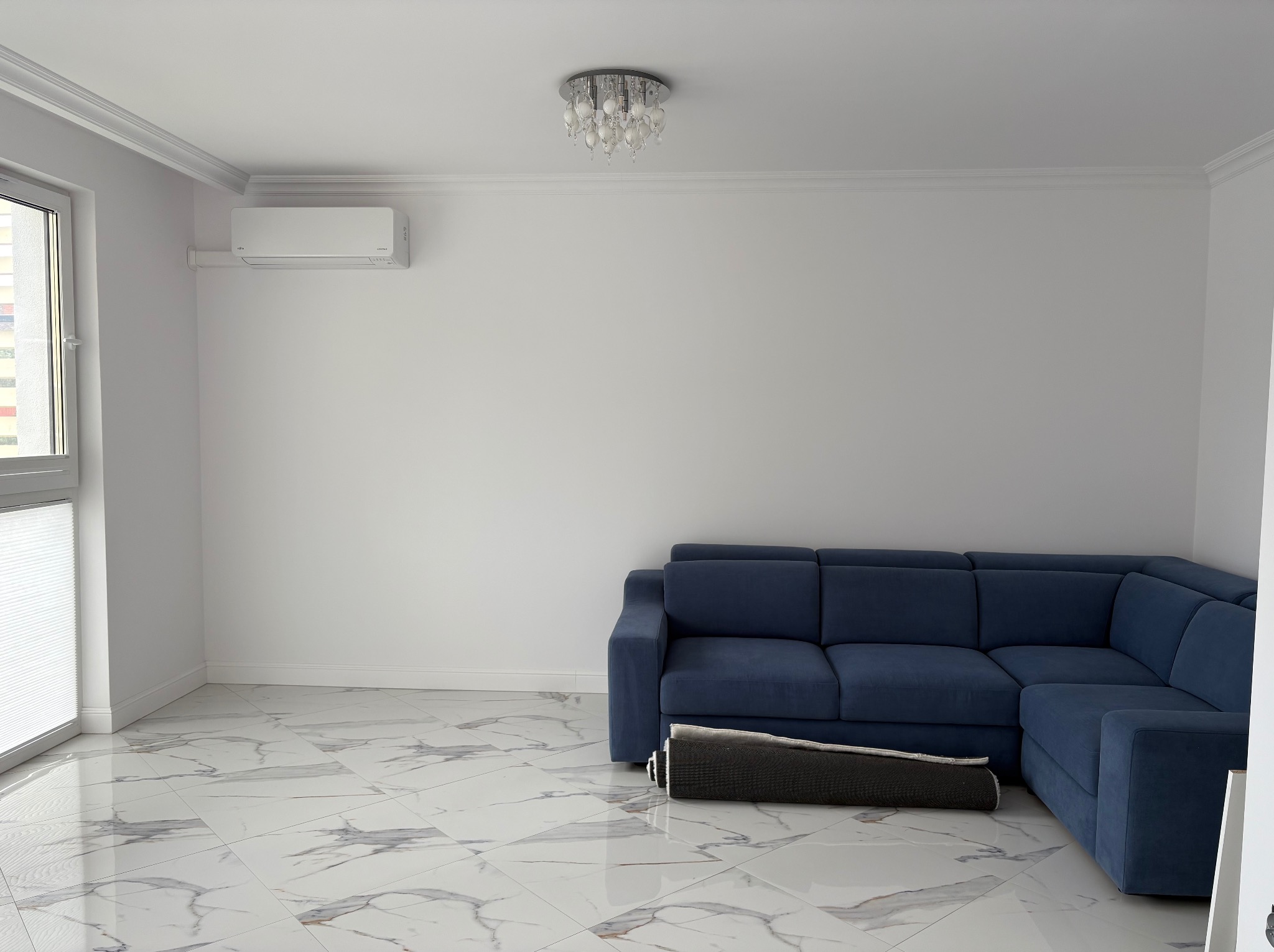 Białe wnętrze z zamontowaną klimatyzacją Fujitsu, niebieskim narożnikiem i marmurową podłogą. Nowoczesny żyrandol sufitowy. Widok na okno.