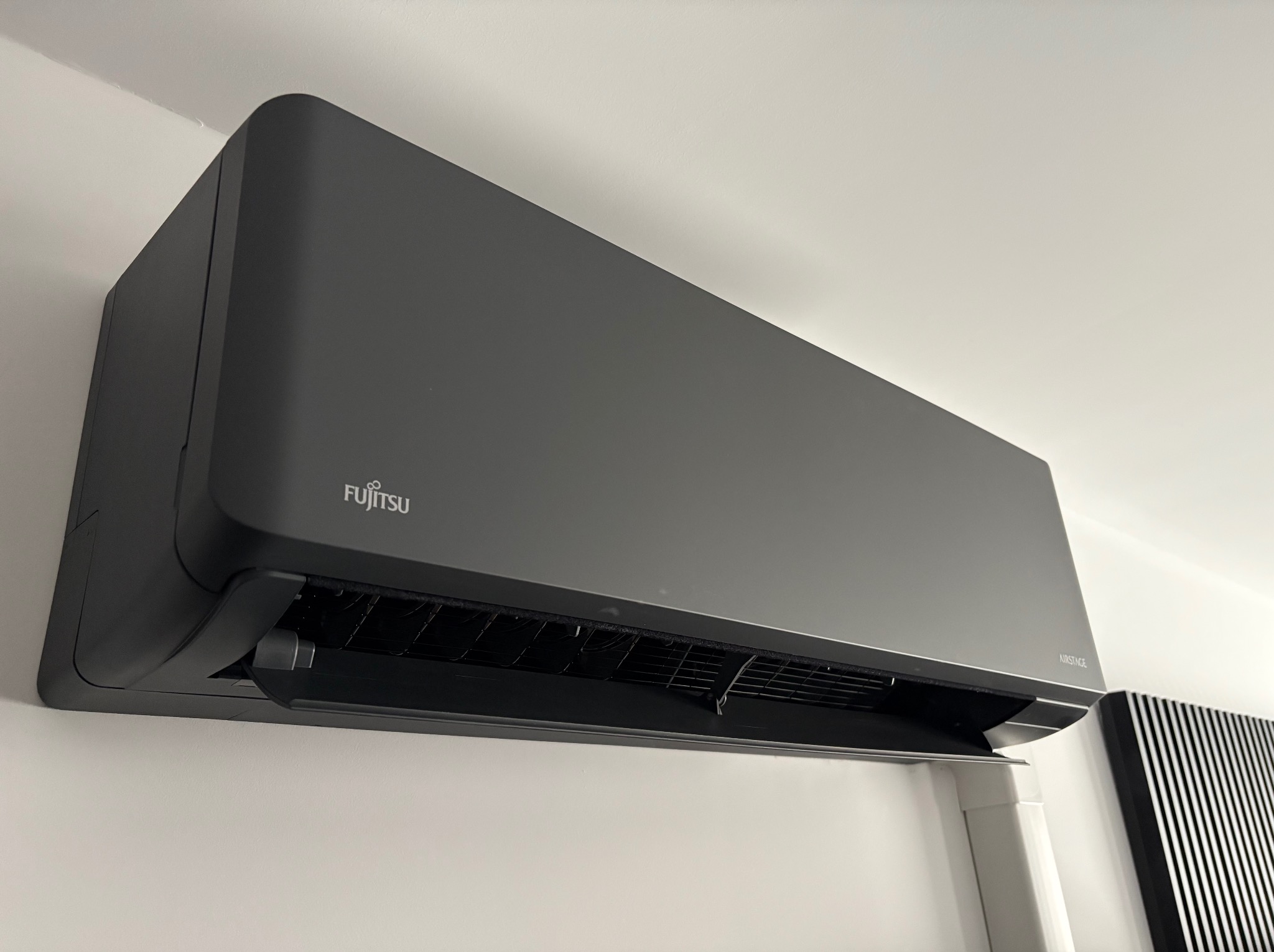 Elegancki, czarny klimatyzator Fujitsu Airstage zamontowany na białej ścianie, widoczny z boku, z otwartą klapą wlotu powietrza. Minimalistyczny design.