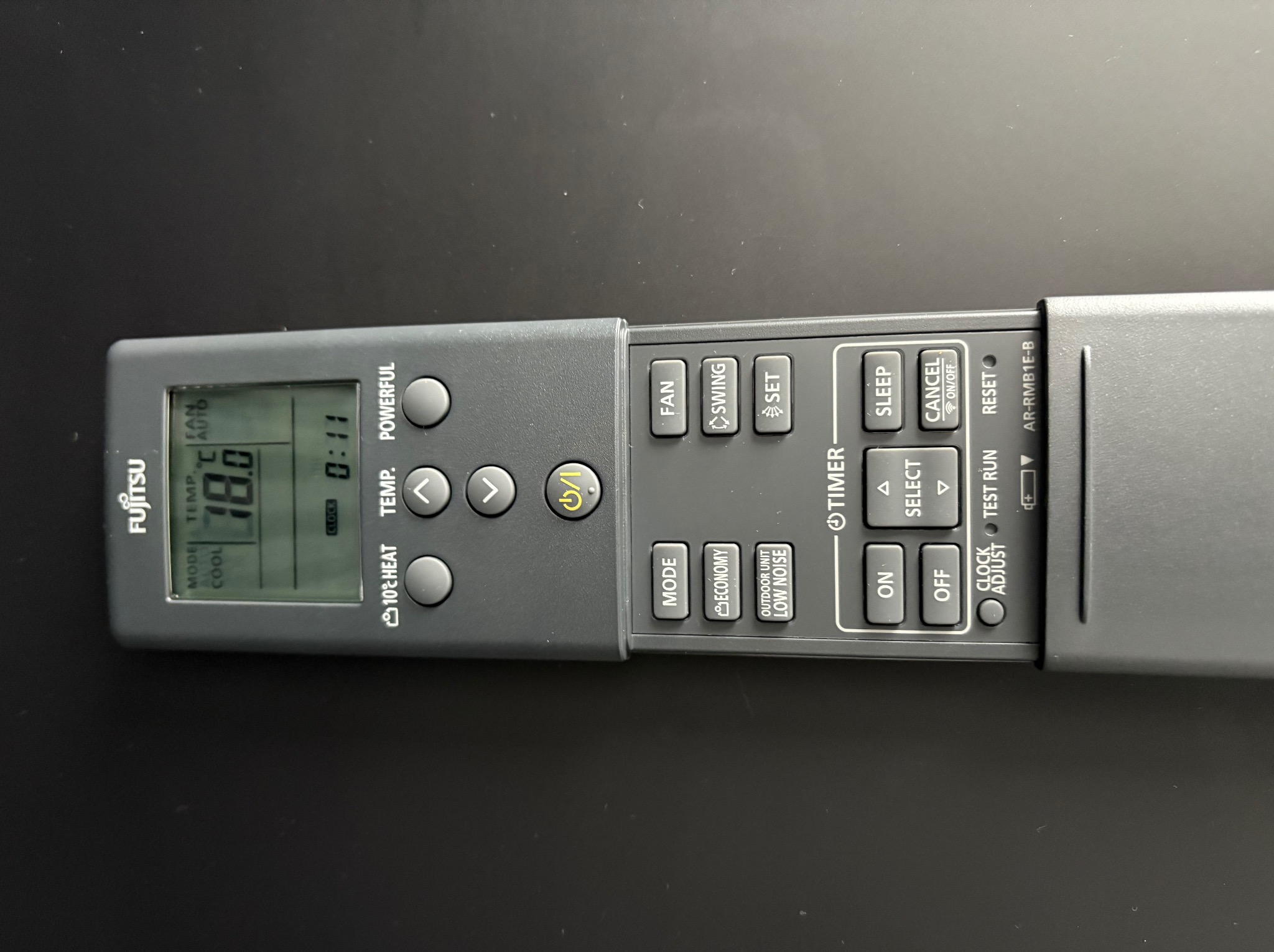 Szary pilot klimatyzacji Fujitsu z wyświetlaczem LCD pokazującym temperaturę 18 stopni i tryb chłodzenia, leżący na czarnym tle. Widoczne przyciski funkcyjne.