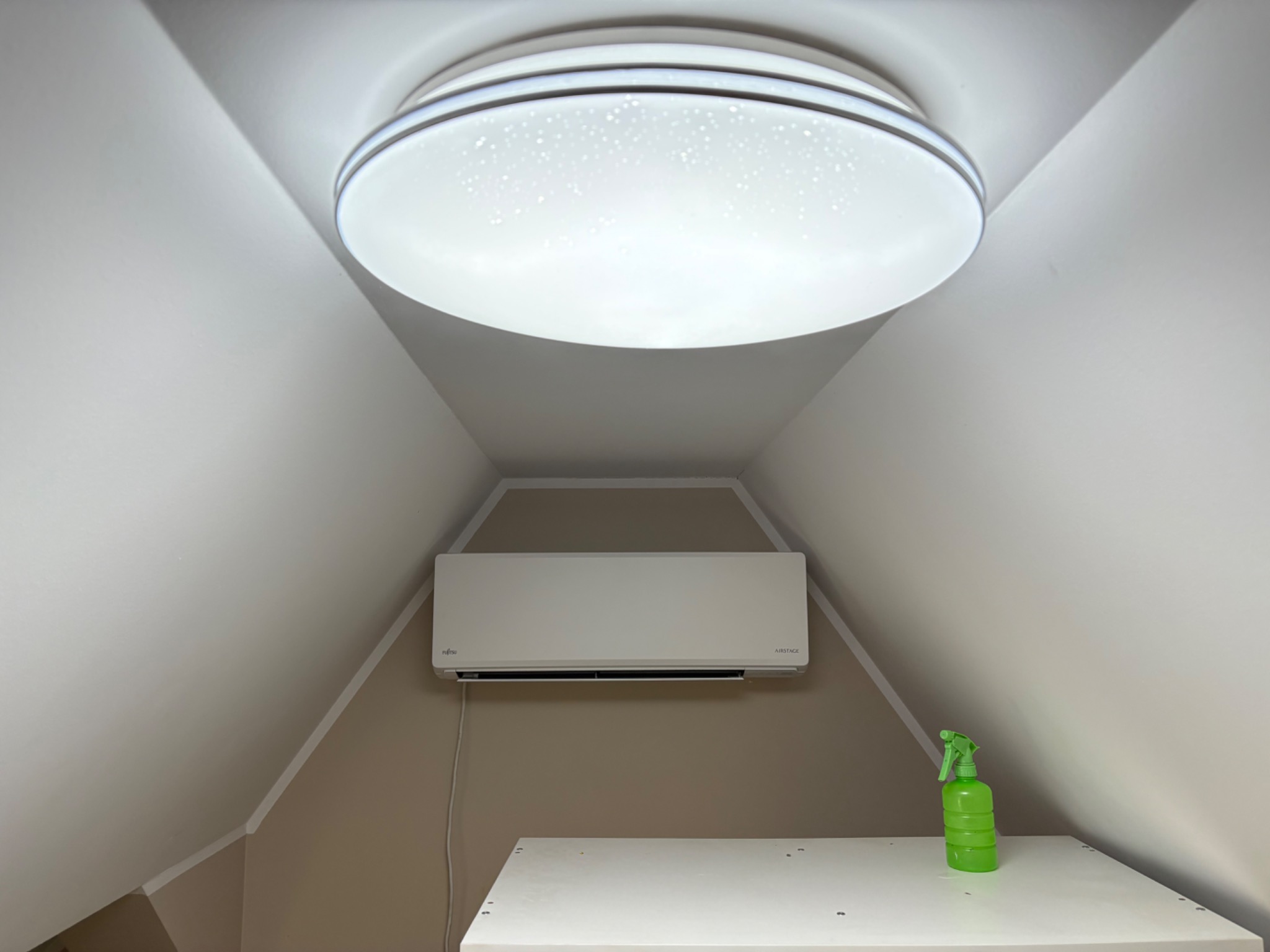 Klimatyzator Fujitsu AIRSTAGE zamontowany pod skośnym sufitem, oświetlony okrągłą lampą sufitową. Na pierwszym planie zielony spryskiwacz na białej powierzchni.