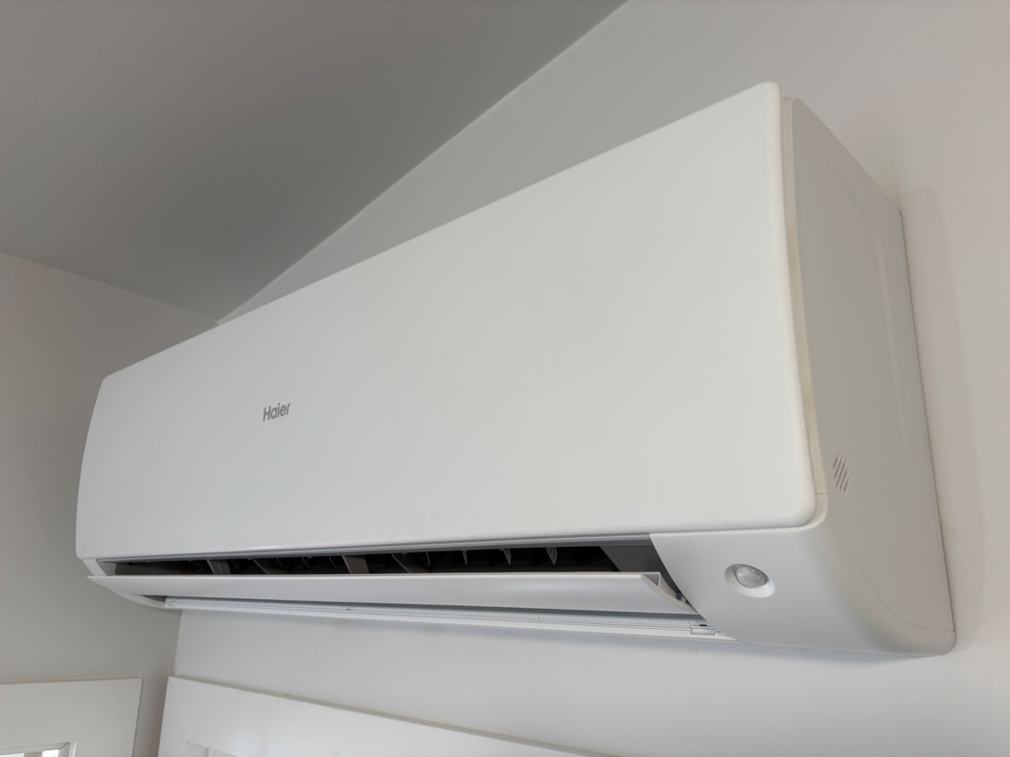 Biała klimatyzacja Haier zamontowana na ścianie w jasnym wnętrzu, widoczna z boku, z otwartą klapą nawiewu. Minimalistyczny design.