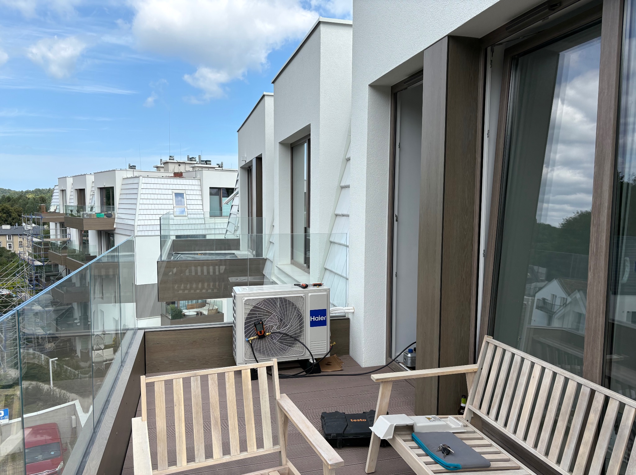 Biała jednostka zewnętrzna klimatyzacji Haier na balkonie apartamentu, narzędzia serwisowe obok, widok na nowoczesną architekturę miejską i błękitne niebo.