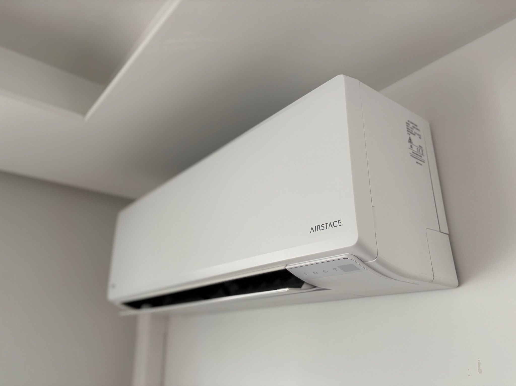 Biała jednostka klimatyzacji AIRSTAGE zamontowana na ścianie w jasnym wnętrzu, widok z dołu, subtelne oświetlenie podkreśla nowoczesny design urządzenia.