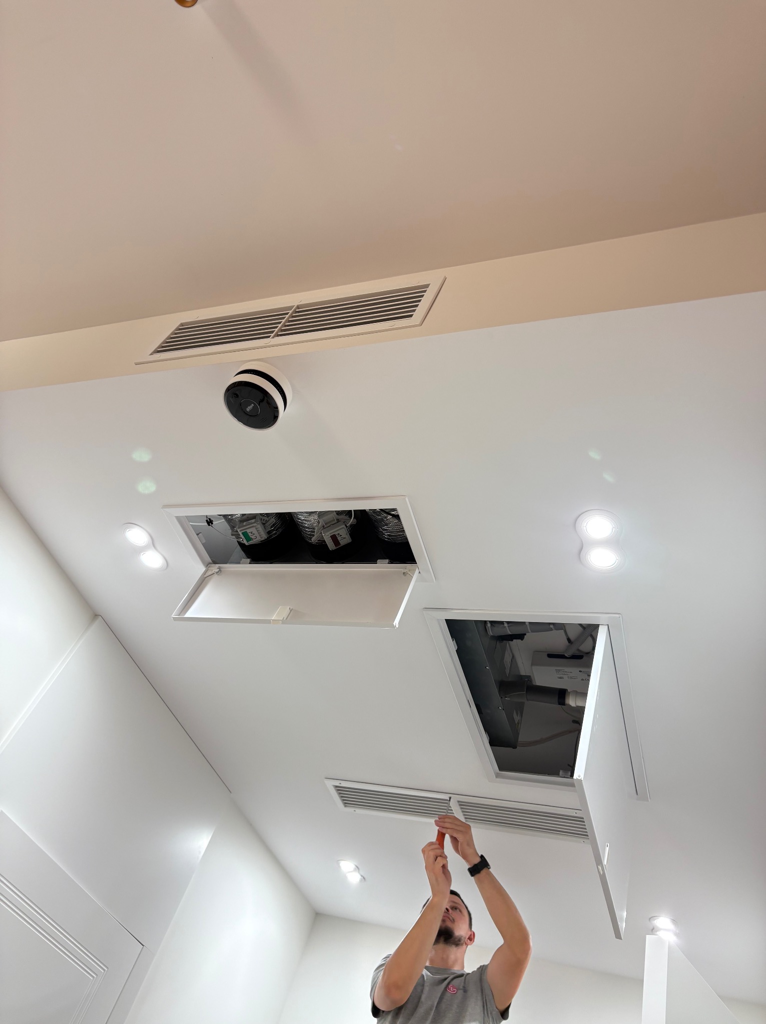 Instalator w trakcie montażu klimatyzacji kanałowej w suficie podwieszanym, widoczne kratki wentylacyjne i otwory rewizyjne. Białe wnętrze, oświetlenie punktowe.