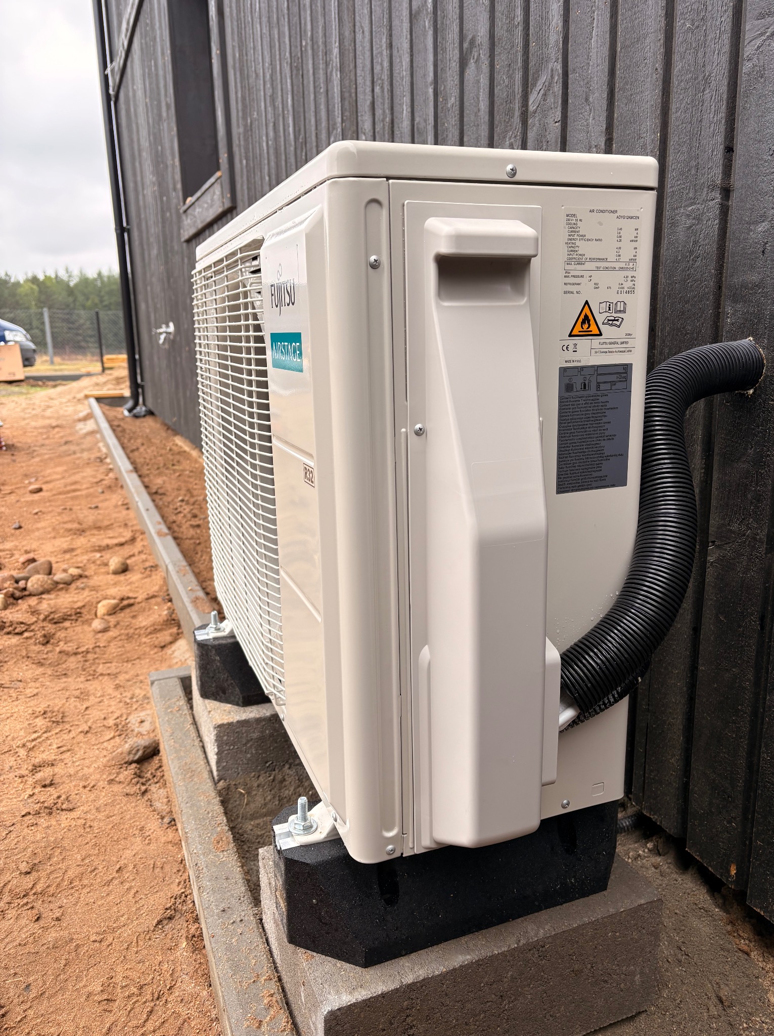 Zewnętrzna jednostka klimatyzacji Fujitsu Airstage zamontowana na betonowych bloczkach przy ciemnej, drewnianej elewacji budynku. Widoczny czarny, karbowany przewód.