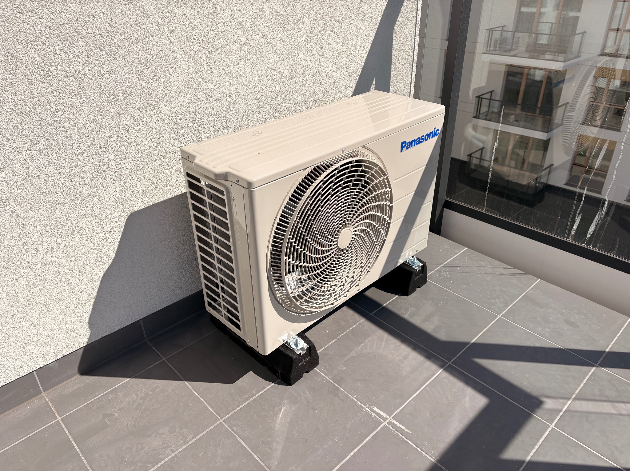 Kompaktowa jednostka zewnętrzna klimatyzacji Panasonic, zamontowana na balkonie z szarymi płytkami, z widocznym fragmentem okna i odbiciem budynku.