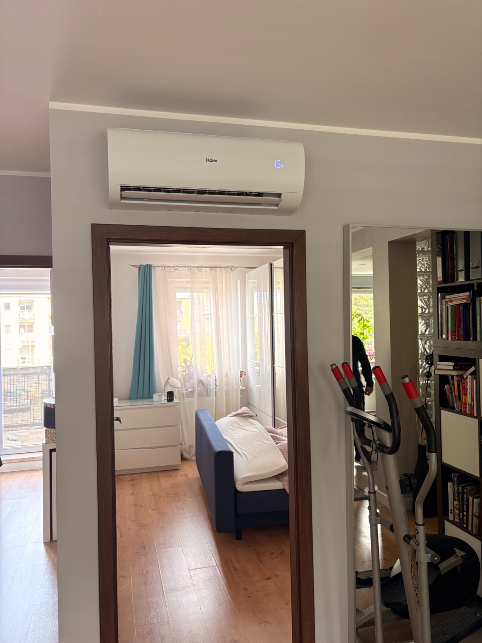 Biały klimatyzator Haier na ścianie w pokoju, widoczny przez futrynę drzwi. W tle sypialnia z niebieską sofą i biurko. Z boku widoczny orbitrek i regał z książkami.