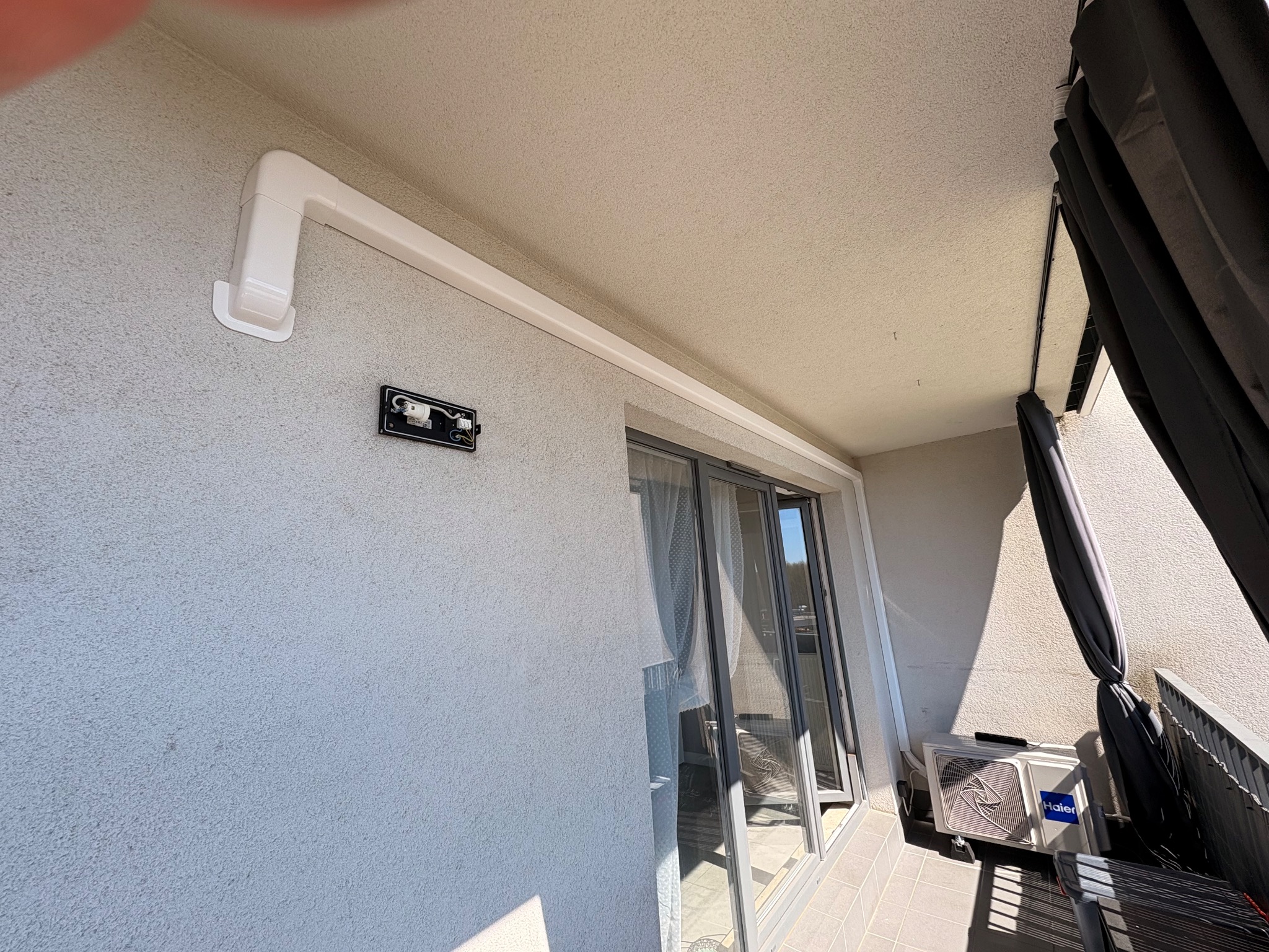 Biała rura instalacyjna klimatyzacji na balkonie, z widoczną jednostką zewnętrzną Haier i fragmentem okna balkonowego. Ściana o szarej, chropowatej fakturze.