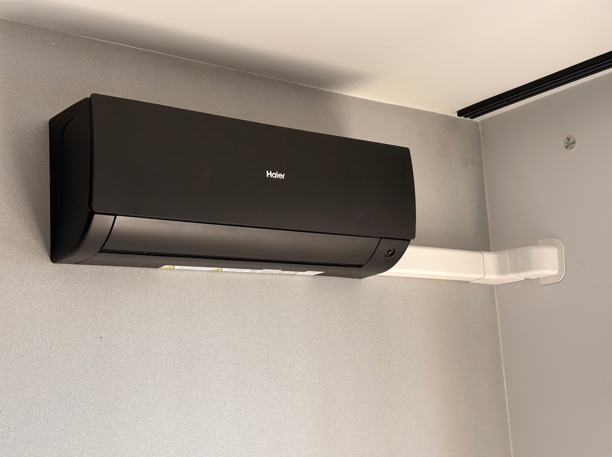 Czarny klimatyzator Haier zamontowany na szarej ścianie, z widoczną białą rurą odprowadzającą skropliny. Minimalistyczny design, nowoczesne wnętrze.