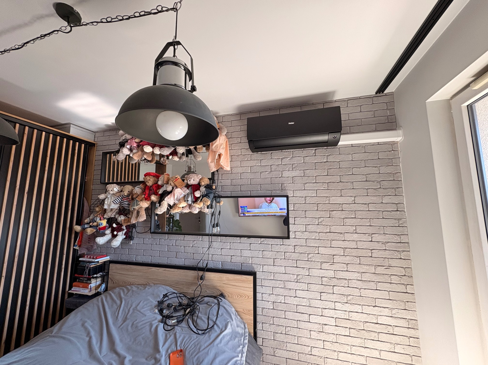 Czarny klimatyzator Haier zamontowany na ceglanej ścianie w sypialni. Nad łóżkiem wisi lustro z telewizorem i lampa w stylu industrialnym. Dekoracje z pluszaków.