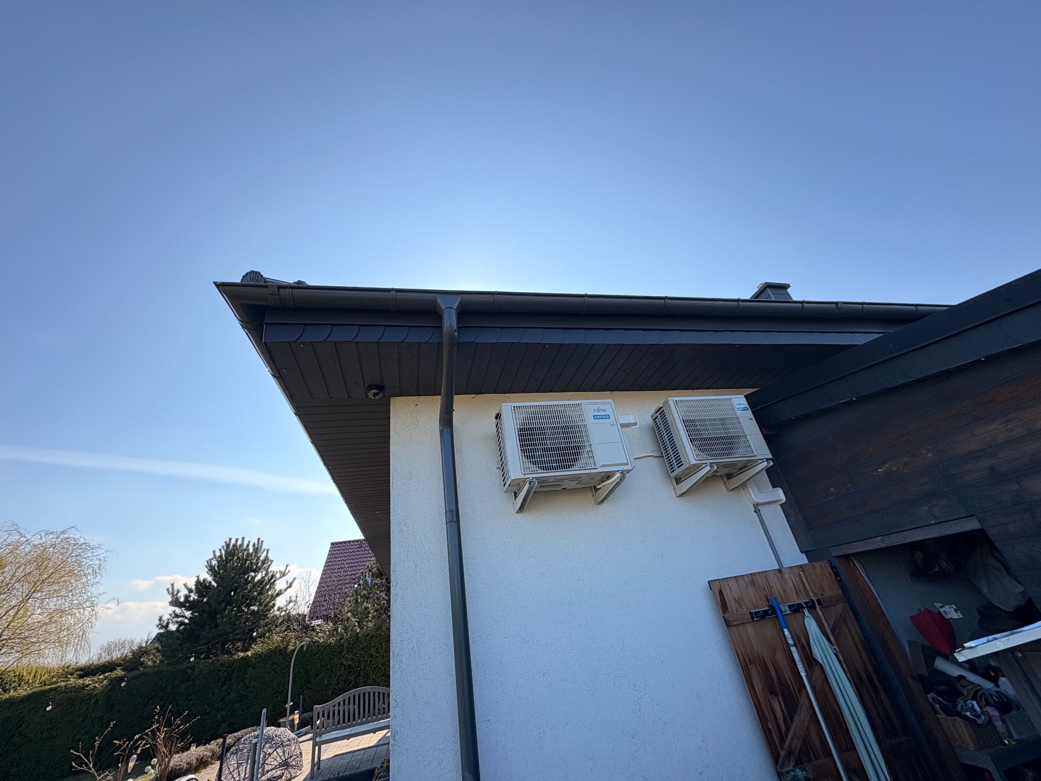 Dwa białe klimatyzatory Fujitsu Airstage zamontowane na elewacji domu, widoczne z dołu pod kątem, z fragmentem dachu i błękitnym niebem w tle.