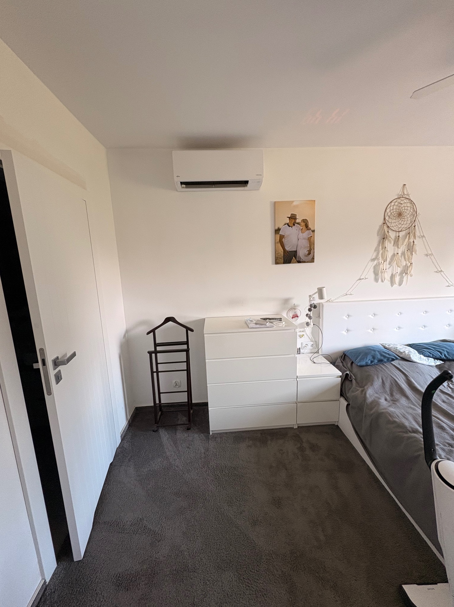 Biała klimatyzacja ścienna w sypialni z szarą wykładziną, widoczna nad komodą. Na ścianie obraz i łapacz snów. Minimalistyczny design wnętrza.