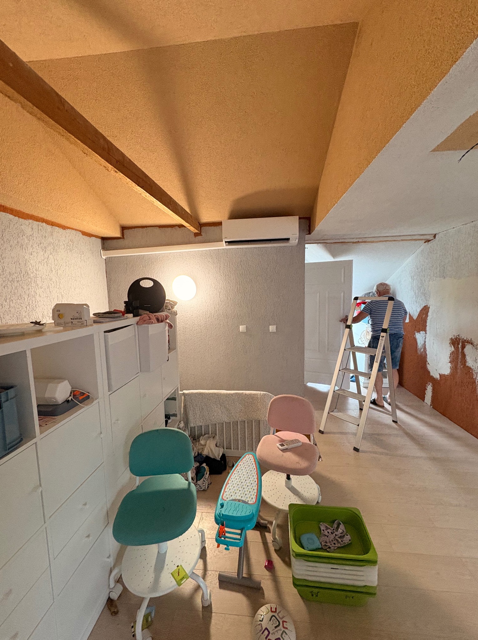 Malowanie ścian w pokoju z klimatyzacją. Mężczyzna na drabinie maluje ścianę na brązowo. Widoczny klimatyzator na ścianie i dziecięce meble.