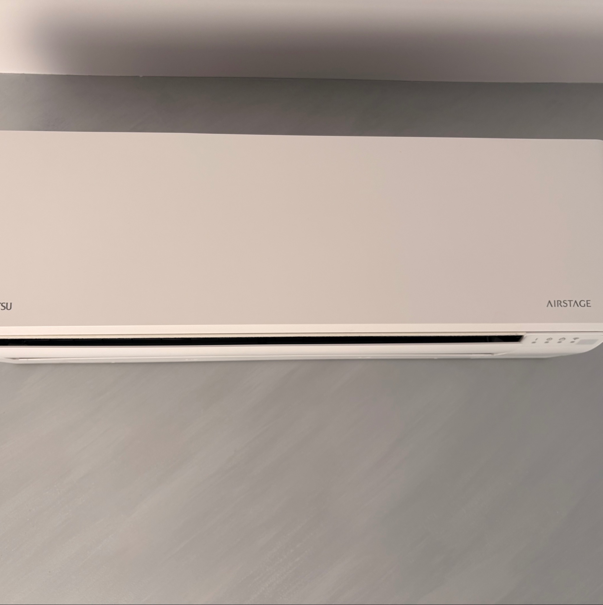 Klimatyzator marki Airstage na szarej ścianie. Widoczne logo producenta. Minimalistyczny design urządzenia. Delikatne oświetlenie podkreśla fakturę ściany.