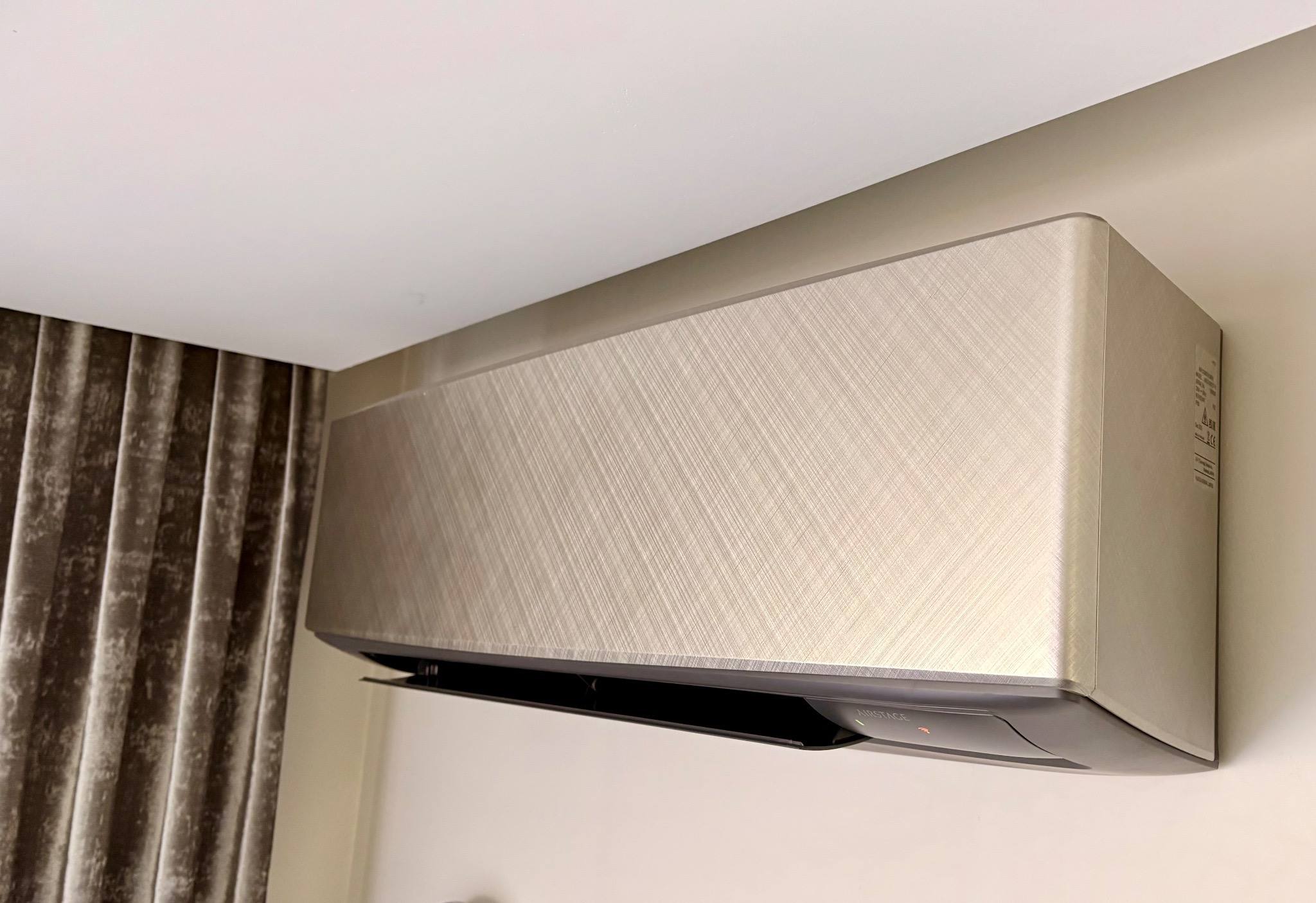 Elegancki klimatyzator ścienny AIRSTAGE o metalicznej, szczotkowanej fakturze, zamontowany w nowoczesnym wnętrzu z zasłonami w tle. Widoczna dioda sygnalizacyjna.