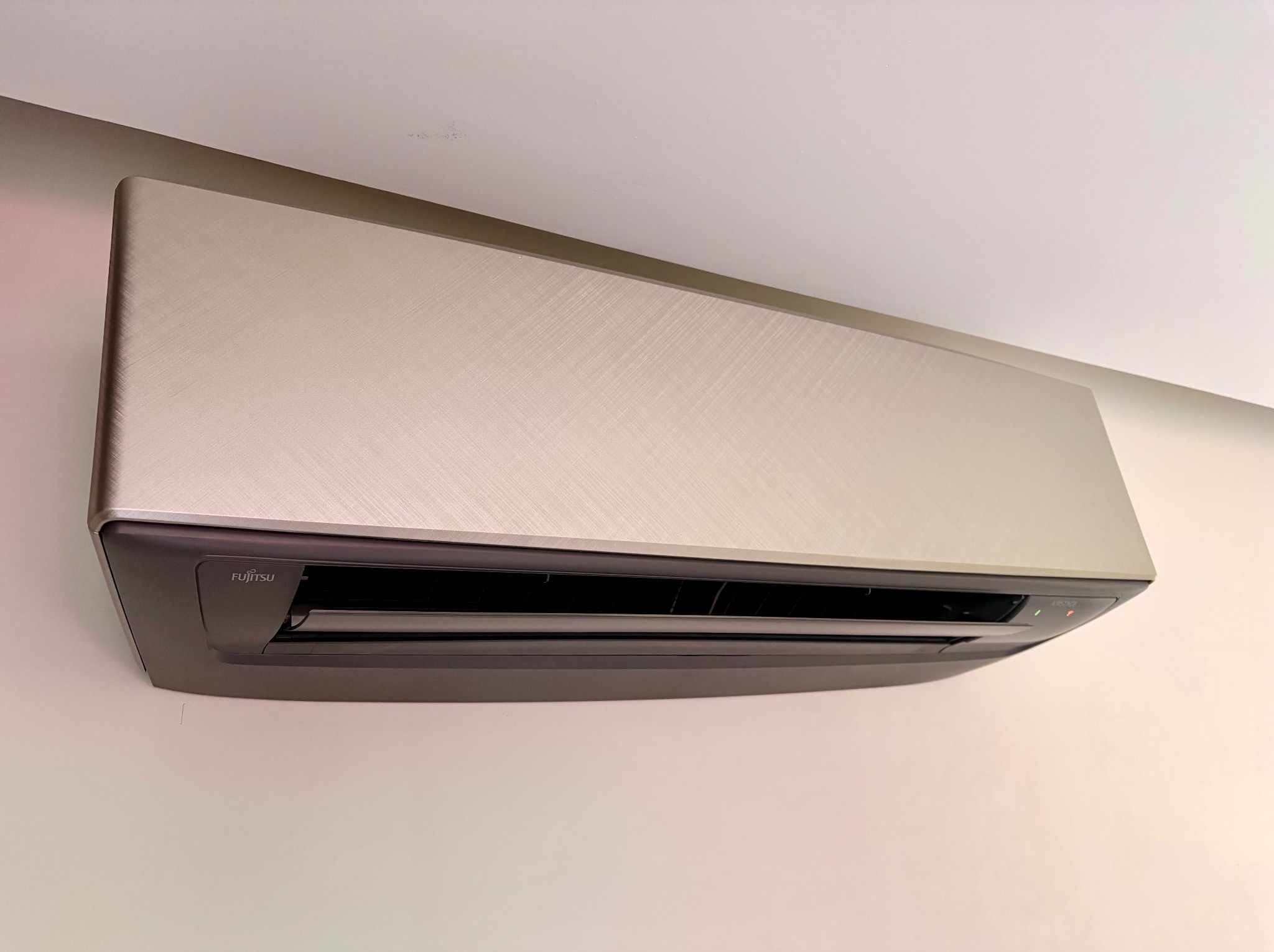 Nowoczesny klimatyzator Fujitsu w kolorze srebrnym, zamontowany na jasnej ścianie. Widoczne detale obudowy i logo producenta. Minimalistyczny design.