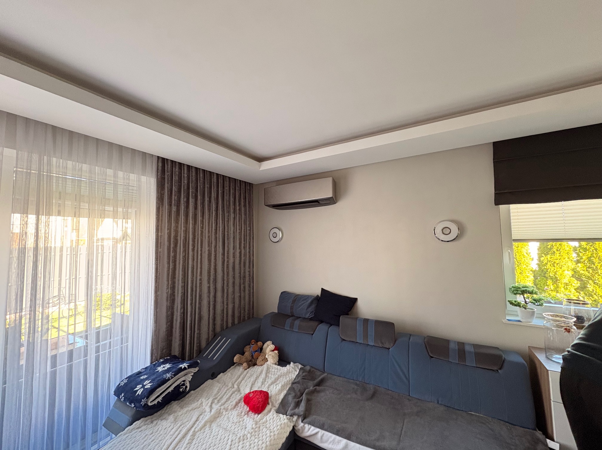 Nowoczesny klimatyzator ścienny w jasnym salonie z dużym oknem i zasłonami. Widoczny fragment sofy i dekoracje. Minimalistyczny design urządzenia.