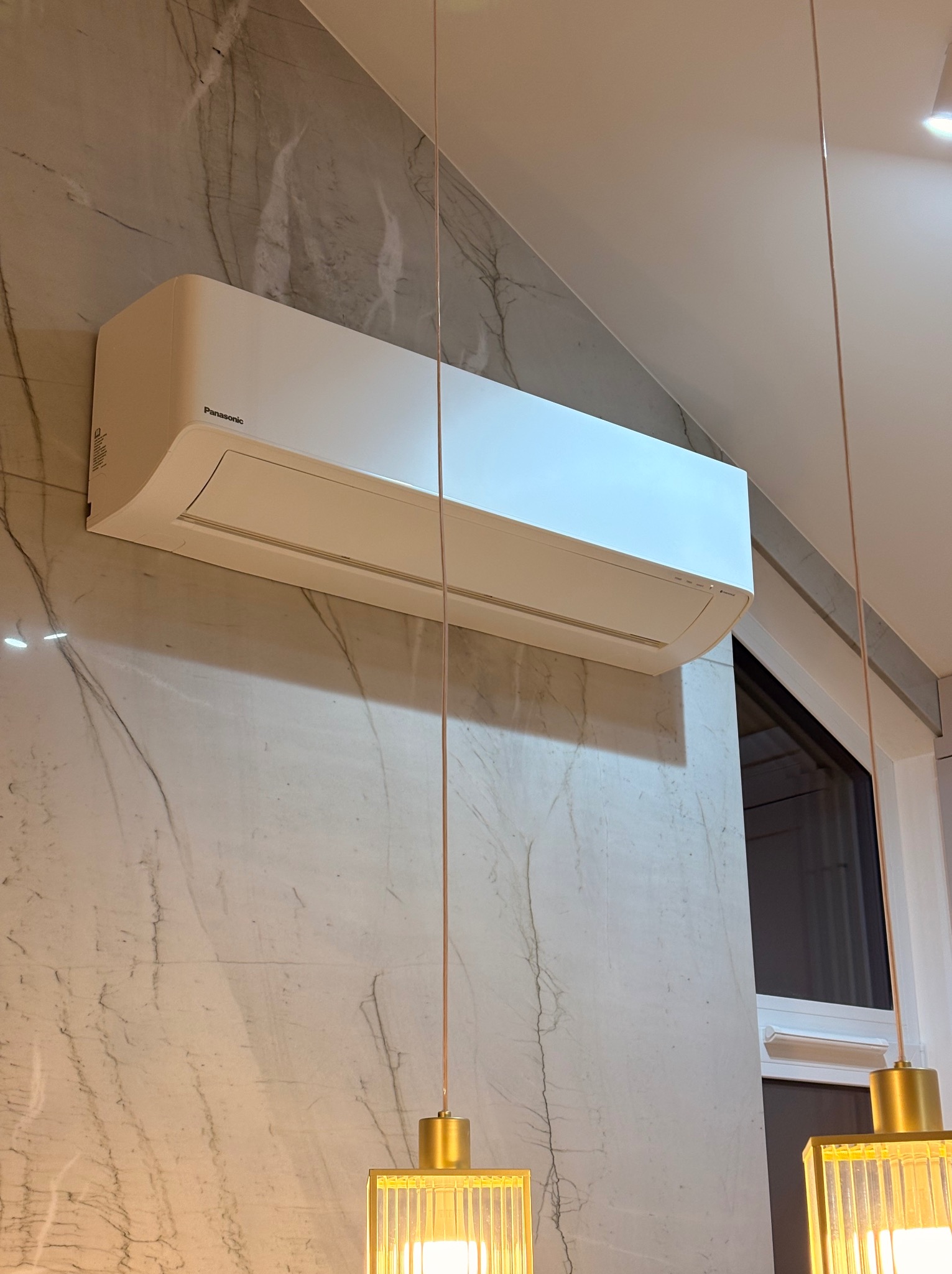 Biała klimatyzacja Panasonic zamontowana na ścianie z marmurowym wzorem, widoczna z dołu, z elementami oświetlenia w kadrze. Nowoczesny design.