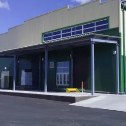 Magazyn z rampą załadunkową, zielono-białe ściany, metalowa konstrukcja zadaszenia i drabina na elewacji.