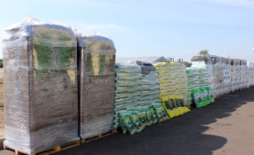 Szeroki kadr z zewnątrz przedstawiający palety z workami ziemi ogrodowej i nawozami, niektóre owinięte folią, stojące na utwardzonym placu w słoneczny dzień.