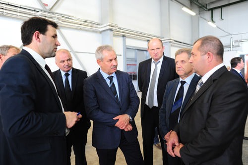 Grupa mężczyzn w garniturach dyskutuje w jasnym, industrialnym wnętrzu, prawdopodobnie podczas spotkania lub inspekcji.