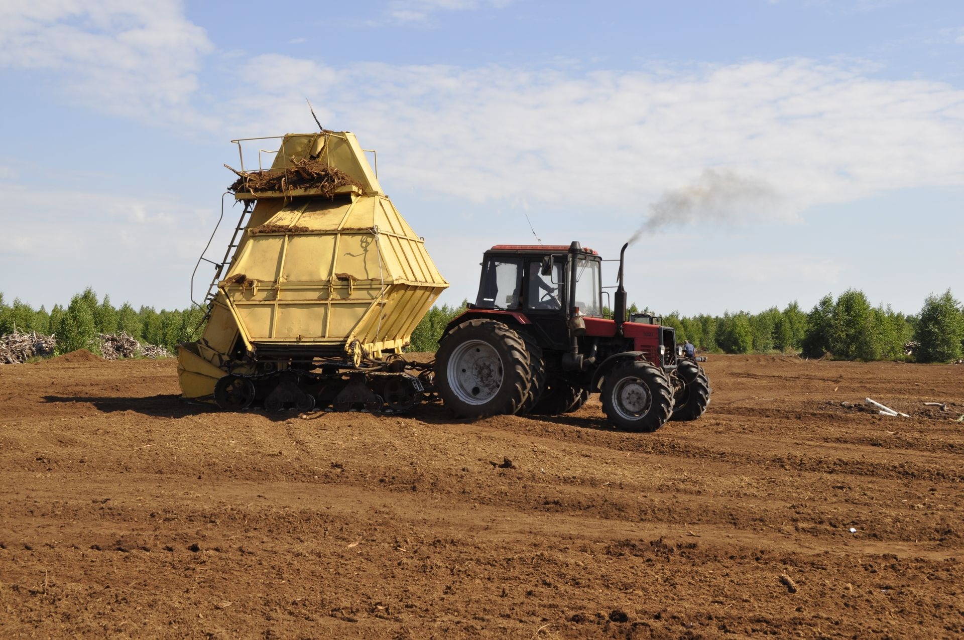 Czerwony traktor z żółtą przyczepą na brązowej, świeżo uprawionej ziemi. W tle widoczne drzewa i błękitne niebo z białymi chmurami.