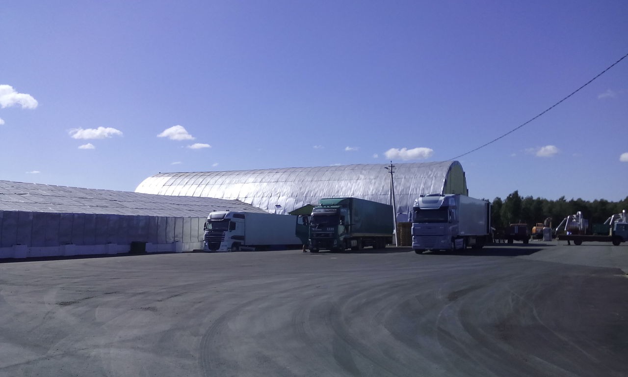 Trzy ciężarówki na placu przed magazynem z nawozami w workach, w tle metalowy hangar i drzewa.