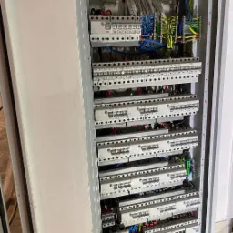 Otwarta skrzynka rozdzielcza z zainstalowanymi bezpiecznikami, przewodami w różnych kolorach i licznikiem energii elektrycznej.