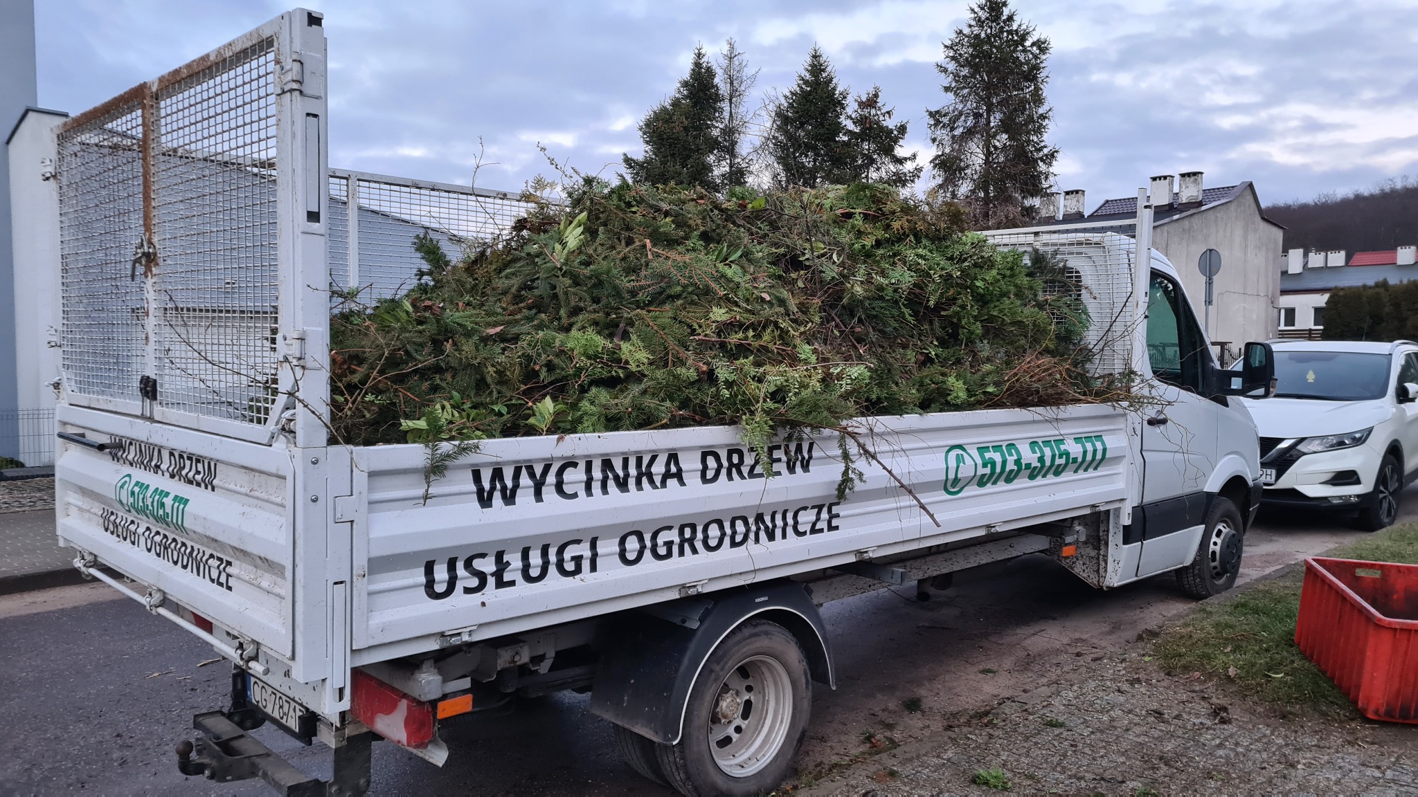 Ciężarówka wypełniona gałęziami i ściętymi drzewami, z widocznym logo firmy ogrodniczej na boku pojazdu, zaparkowana na ulicy w pochmurny dzień.