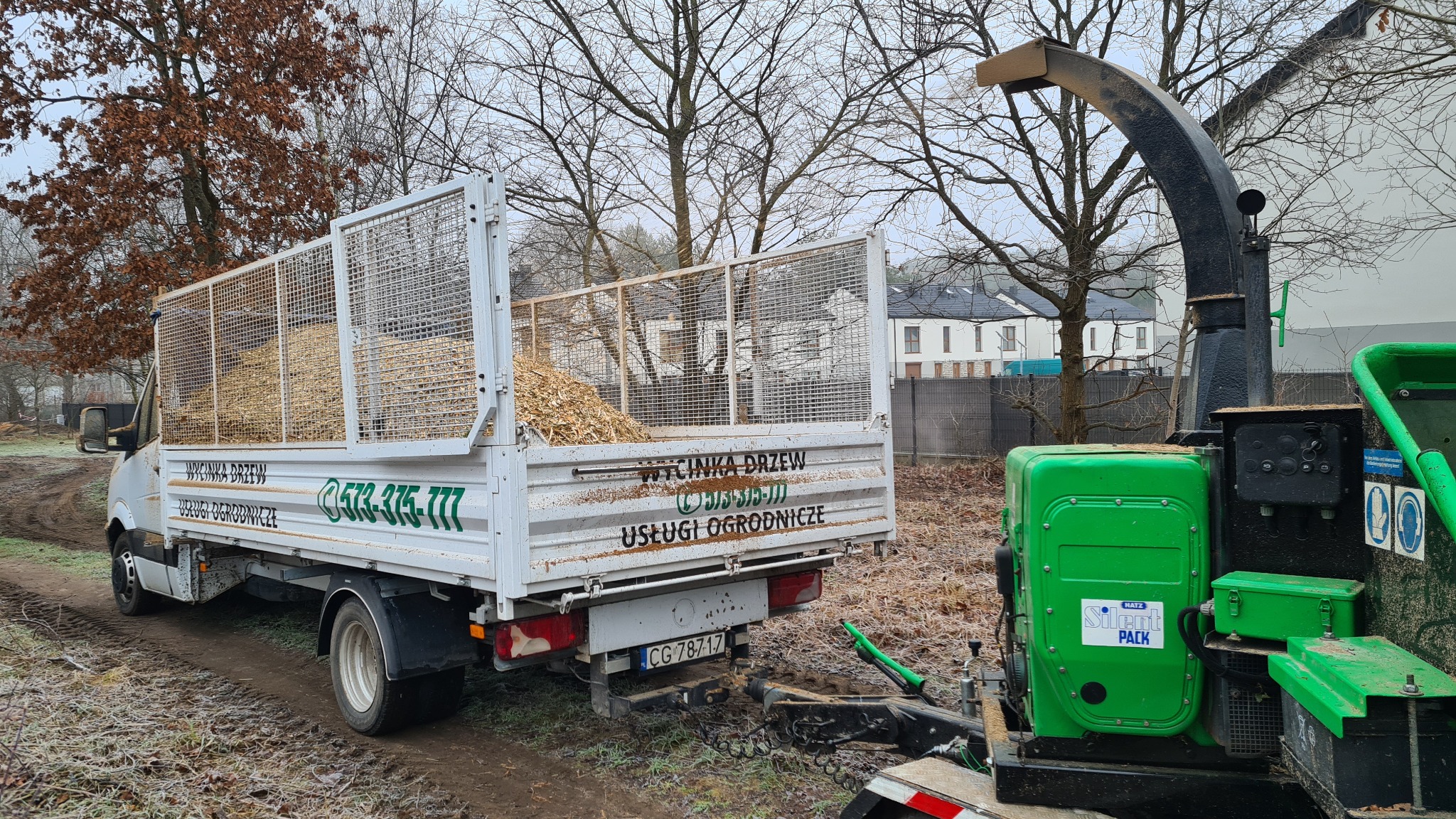 Ciężarówka z przyczepionym rębakiem wypełniona zrębkami drzewnymi stoi na błotnistej drodze w pochmurny dzień, widoczne reklamy usług ogrodniczych na boku pojazdu.