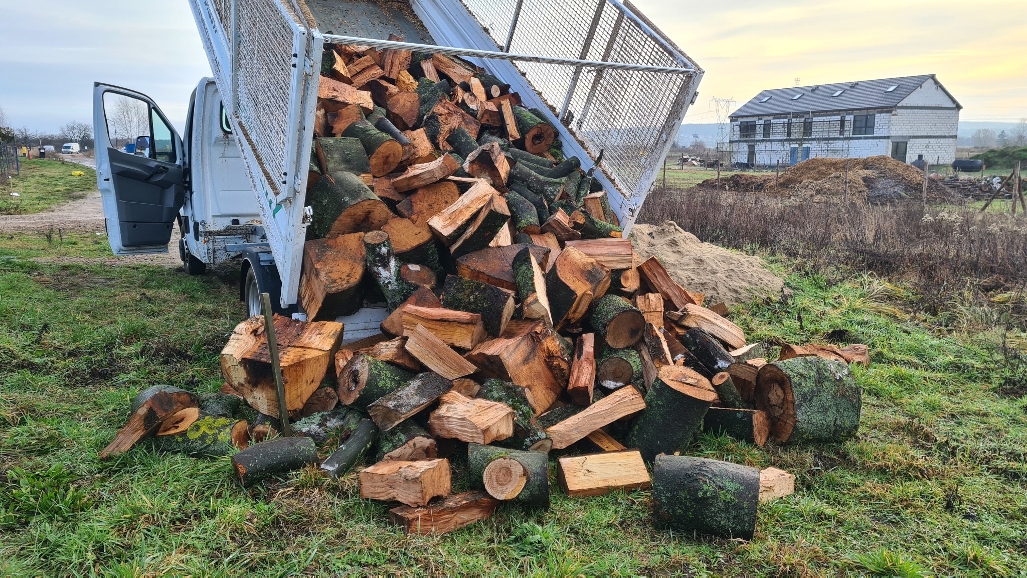 Wyładunek pociętego drewna z wywrotki na trawiastym terenie, w tle budynek w trakcie budowy.