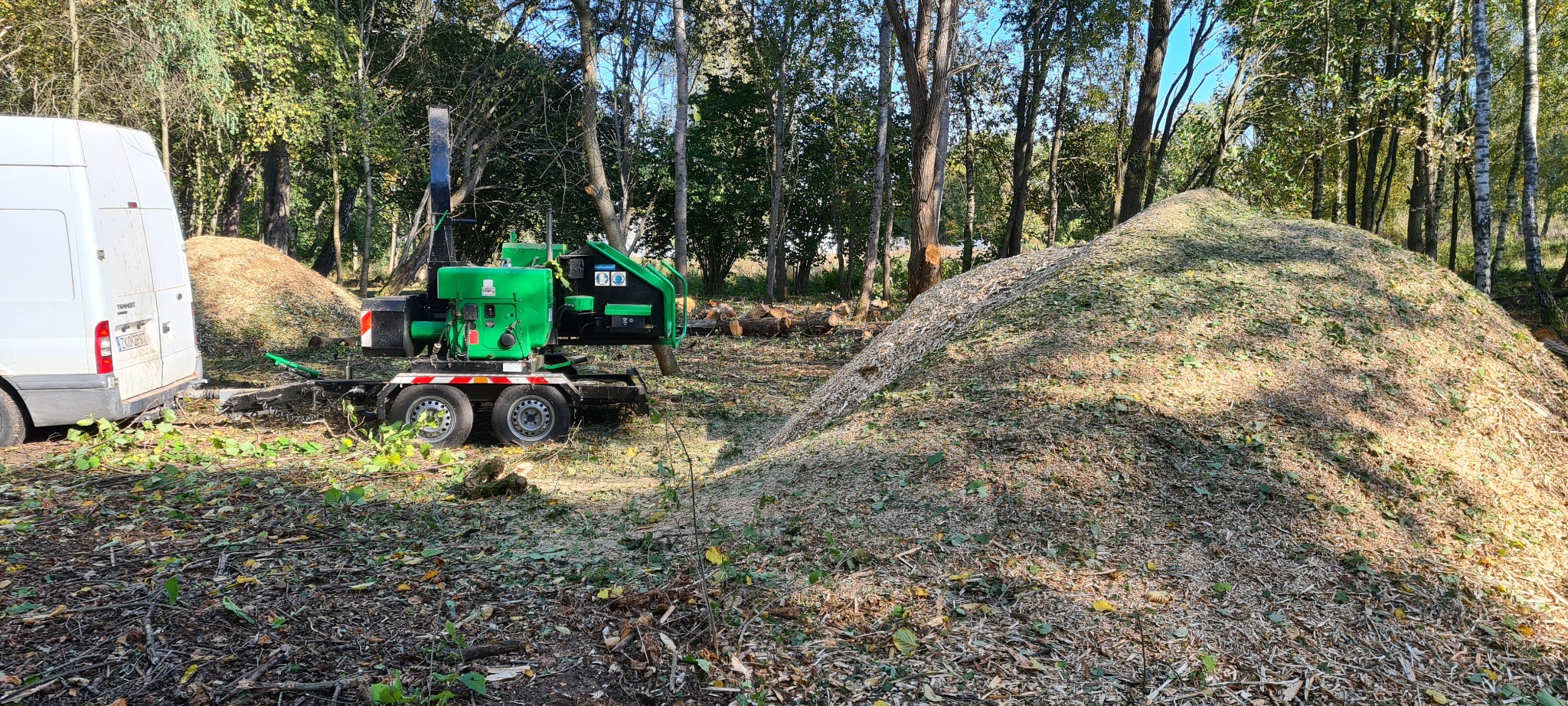 Zielona rębarka do gałęzi przyczepiona do białego vana, obok duży kopiec zrębków drzewnych na tle lasu z powycinanymi pniami drzew.