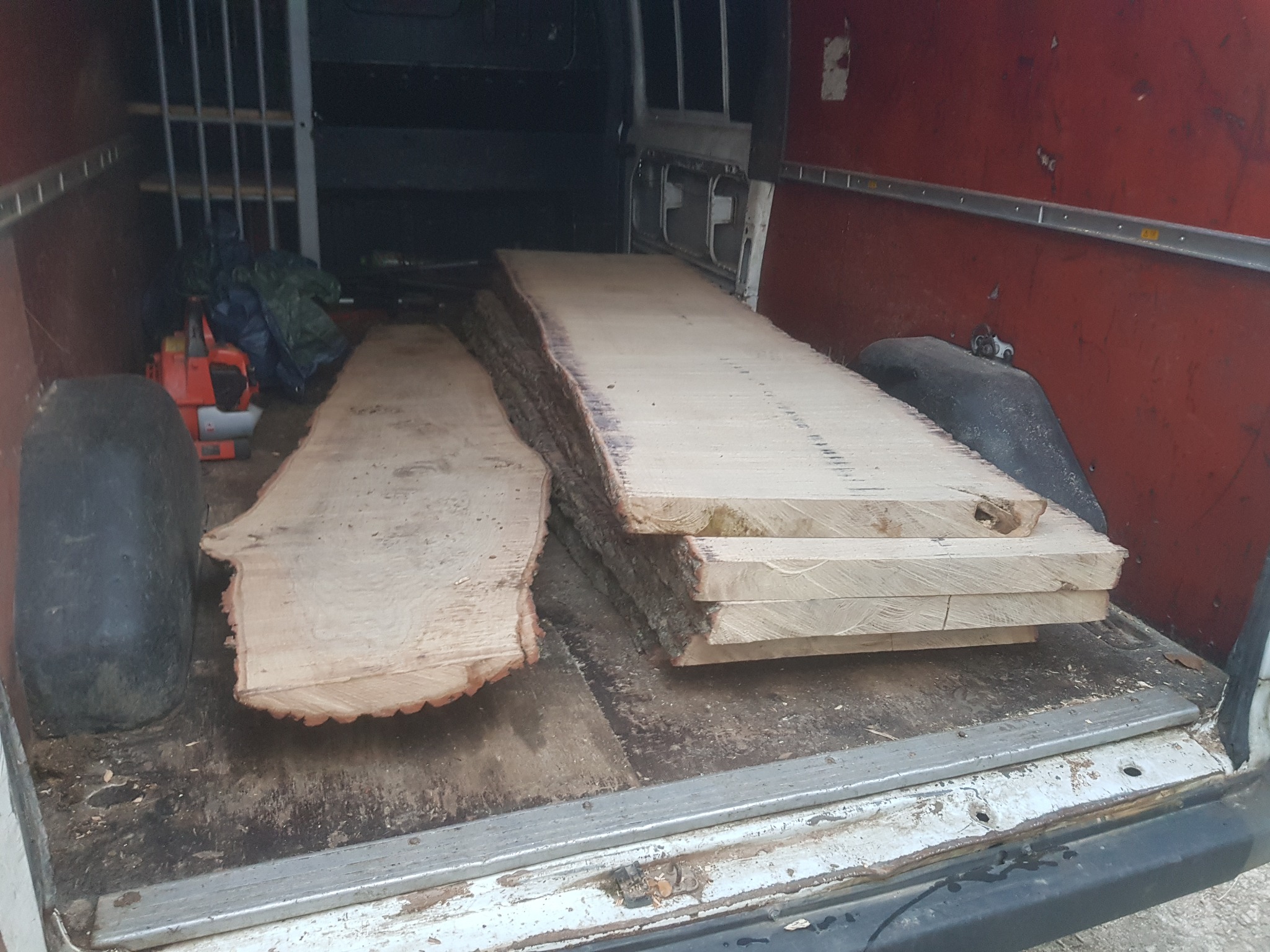 Kawałki drewna z korą załadowane do brudnego busa z piłą spalinową w tle.