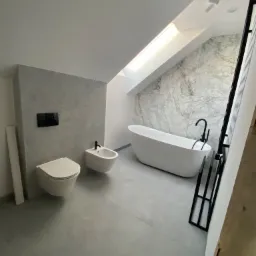 Nowoczesna łazienka na poddaszu ze skośnym oknem dachowym, marmurową ścianą, wolnostojącą białą wanną z czarną armaturą, podwieszaną toaletą i bidetem na tle ściany o betonowej fakturze.