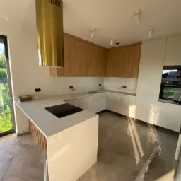 Nowoczesna kuchnia w Zaborowie z białymi i drewnianymi szafkami, złotym okapem cylindrycznym i wyspą z płytą indukcyjną, w trakcie montażu, widoczne przewody elektryczne.