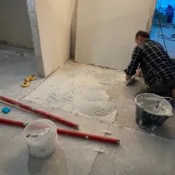 Układanie płytek podłogowych w łazience: glazurnik nanosi klej pacą z zębami na podłogę, widoczne poziomice, przyssawka do płytek i wiadro z zaprawą.