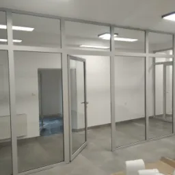 Montaż ścianki działowej z aluminiowymi profilami i przeszklonymi panelami, widoczna otwarta przestrzeń biurowa i sprzęt budowlany.