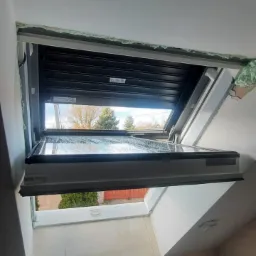 Okno dachowe z roletą zewnętrzną częściowo opuszczoną, widok na zewnątrz z widocznymi drzewami, wykończenie wokół okna w trakcie montażu, widoczna piana montażowa.