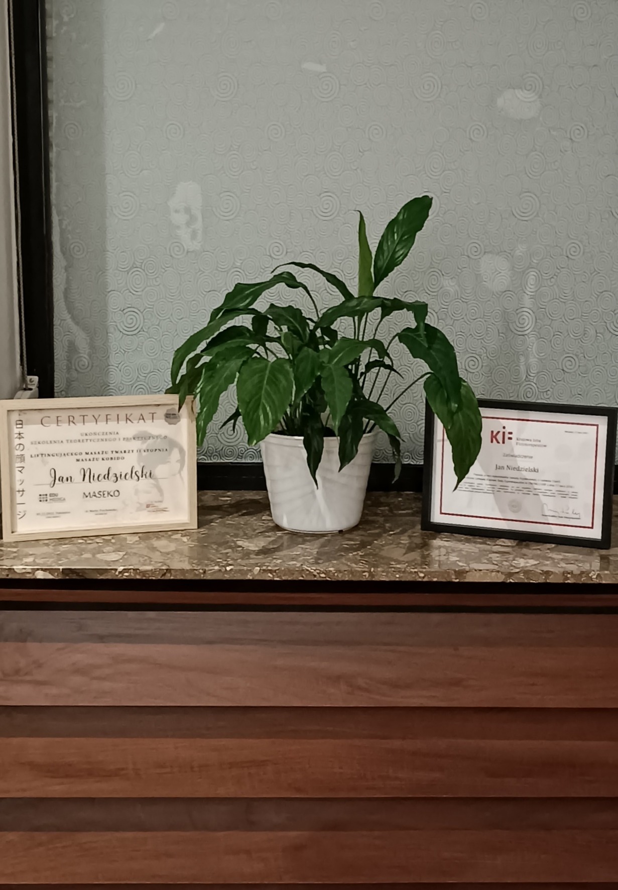 Dwa certyfikaty ukończenia kursów i roślina doniczkowa na blacie w gabinecie, jeden certyfikat w drewnianej ramce dotyczy liftingującego masażu twarzy I stopnia, drugi w czarnej ramce.